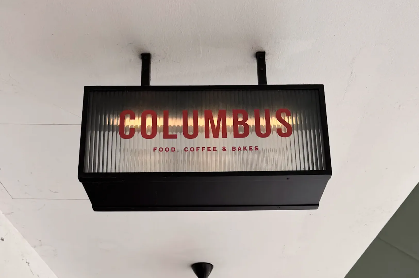 A sleek, modern illuminated rectangular hanging signboard for 'COLUMBUS FOOD, COFFEE & BAKES' with prominent red lettering against a ribbed glass texture under a white ceiling.