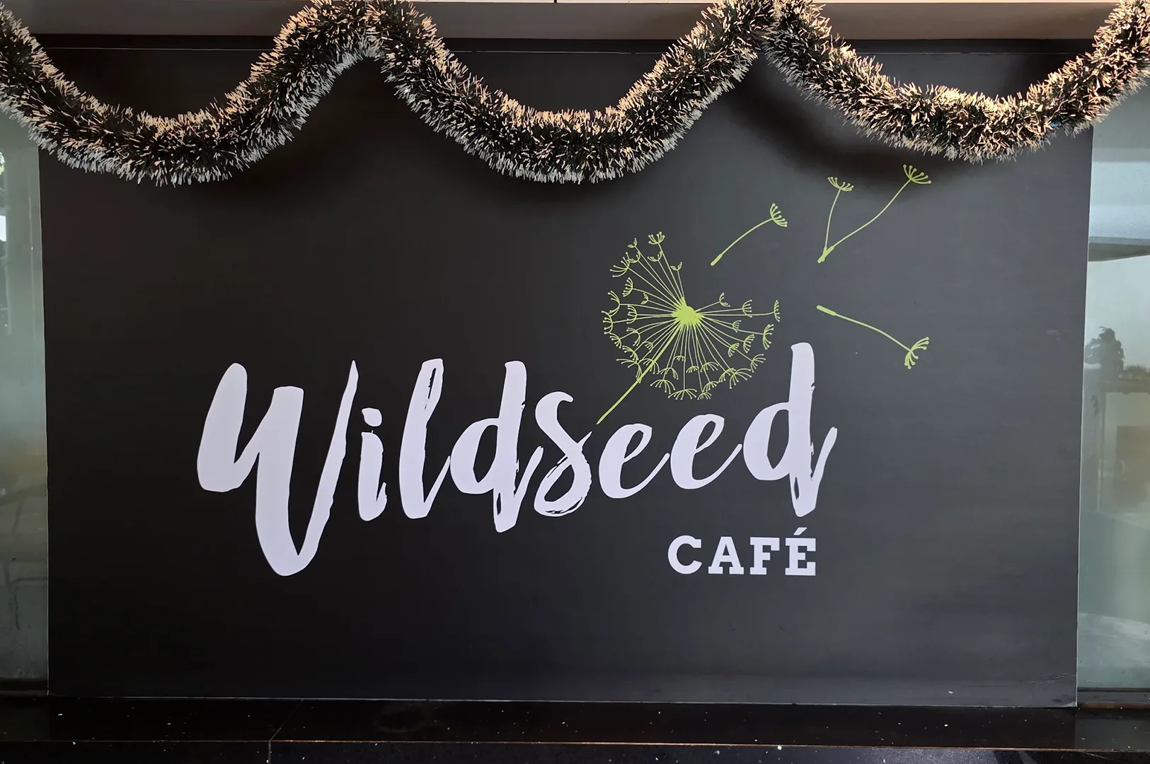 Chalkboard sign featuring the white cursive logo of Wildseed Cafe with a green dandelion graphic, decorated with silver Christmas tinsel garland above.
