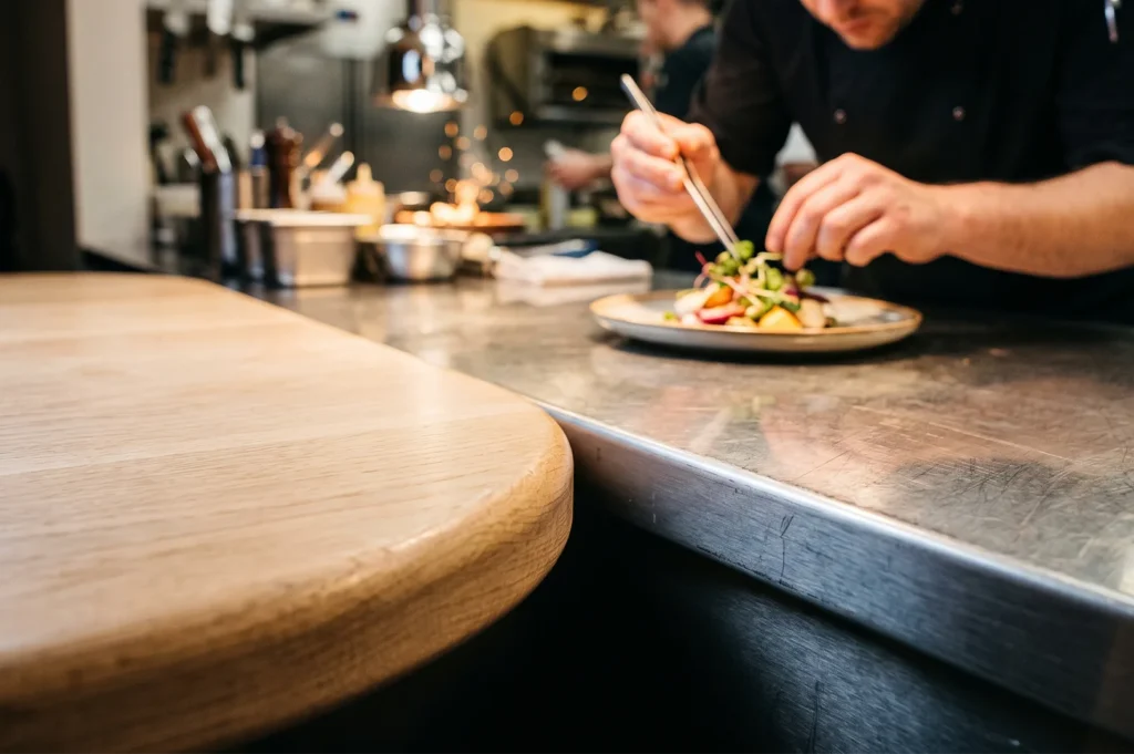 Close-up of a professional chef using precision tweezers to plate a vibrant gourmet salad on a ceramic plate in a commercial restaurant kitchen.