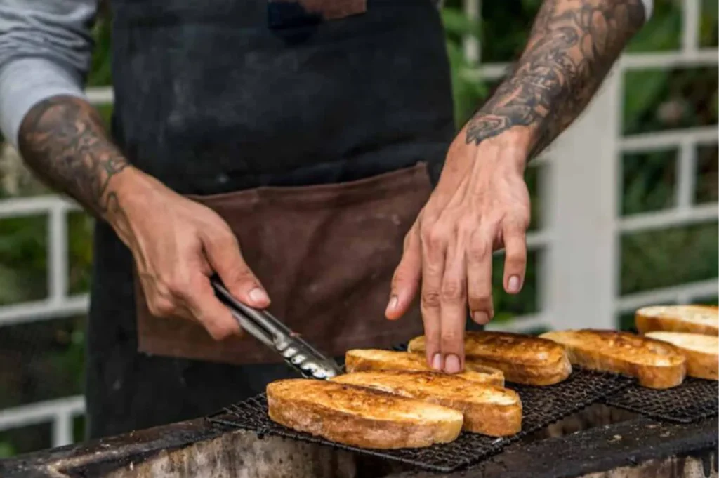 Close-up of a tattooed chef grilling marinated meat skewers and corn on an open flame charcoal grill outdoors.