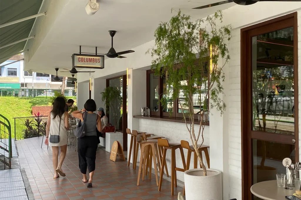 The spacious outdoor patio and walkway of Columbus cafe featuring light wood bar stools, white brick walls, potted green trees, and customers walking past the shopfront.