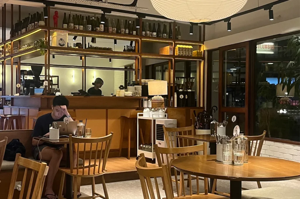 Warm and inviting interior of Columbus cafe with wooden dining tables and chairs, a student working on a laptop, a well-lit service bar with a glass display shelf holding bottles.