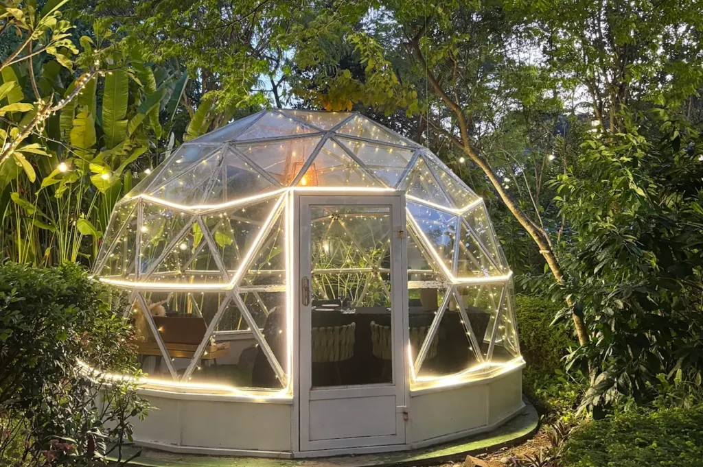An illuminated glass dining dome nestled in a tropical garden at night, decorated with warm LED strip lights and string lights, featuring an intimate table setup inside.