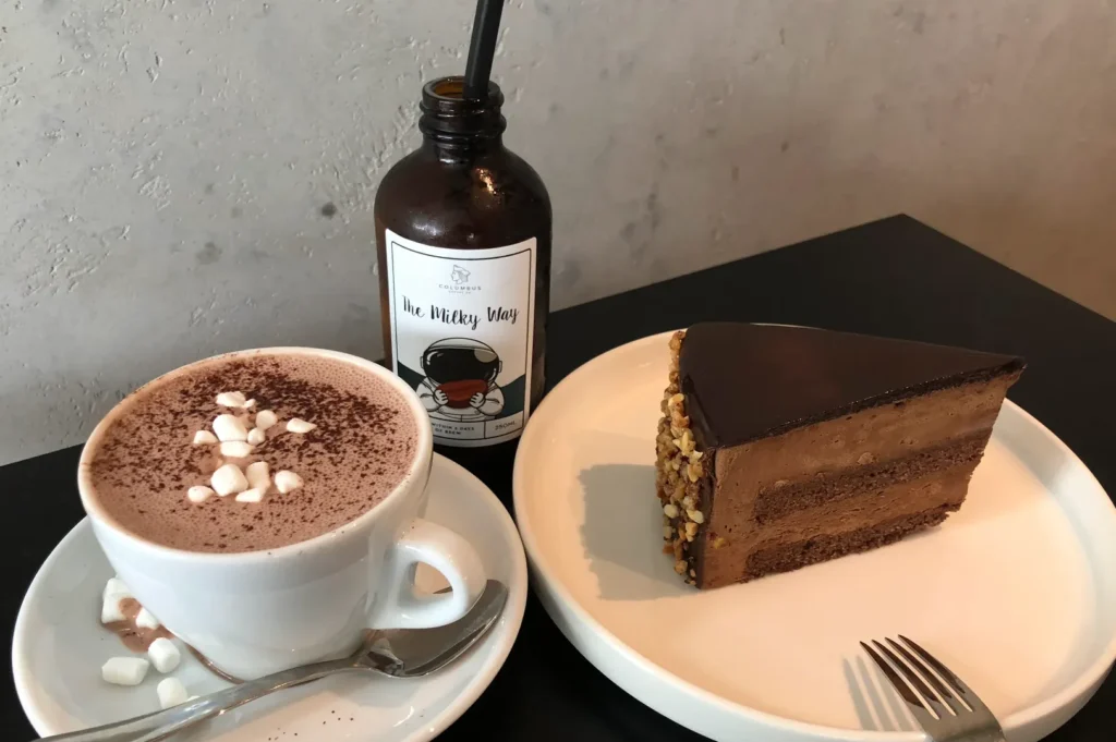 A delicious slice of multi-layered chocolate mousse cake with nut toppings next to a hot cocoa cup with marshmallows and a cold bottled cold brew labeled 'The Milky Way'.