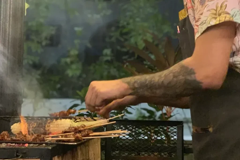A chef using tongs to flip chicken wings on a hot barbecue grill, surrounded by skewers of meat cooking over charcoal.