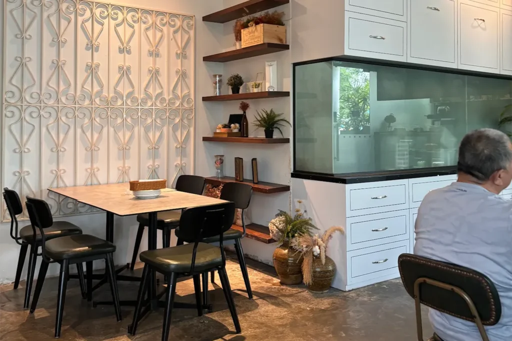 Cozy indoor restaurant dining space with marble-top tables, black industrial chairs, floating wooden shelves filled with plants, and white cabinet storage units.