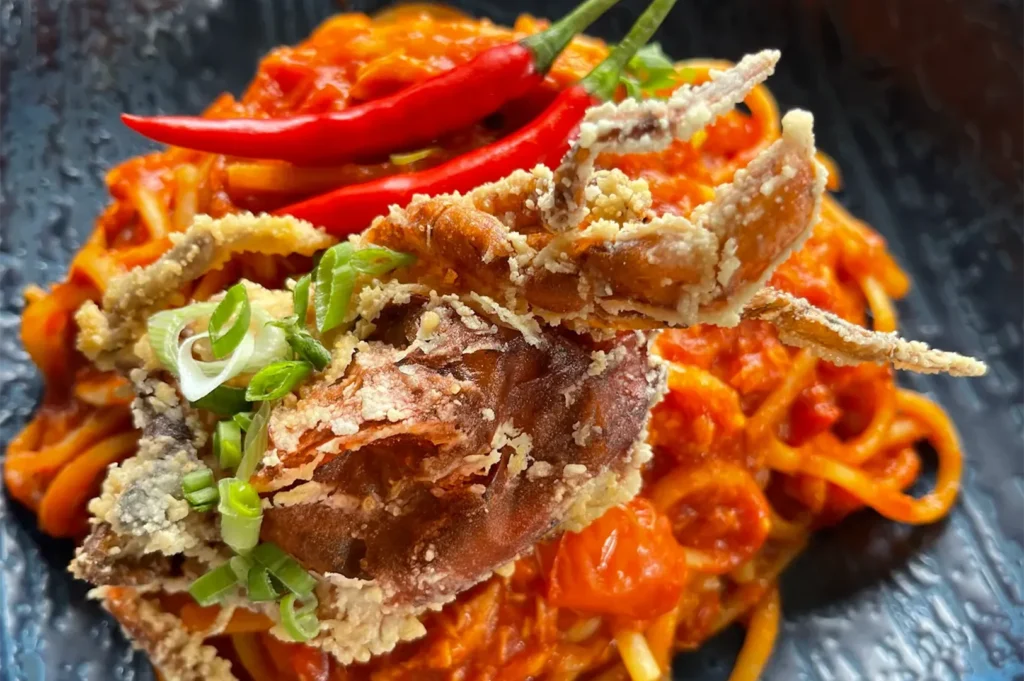 Close-up of appetizing spaghetti pasta tossed in savory spicy tomato chili sauce, garnished with deep-fried soft shell crab, green scallions, and two red chili peppers on top.
