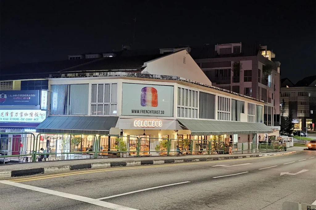 Night view of the Columbus cafe storefront located at a street corner building in Singapore, showcasing a brightly lit interior, green awnings, and surrounding street pavement.