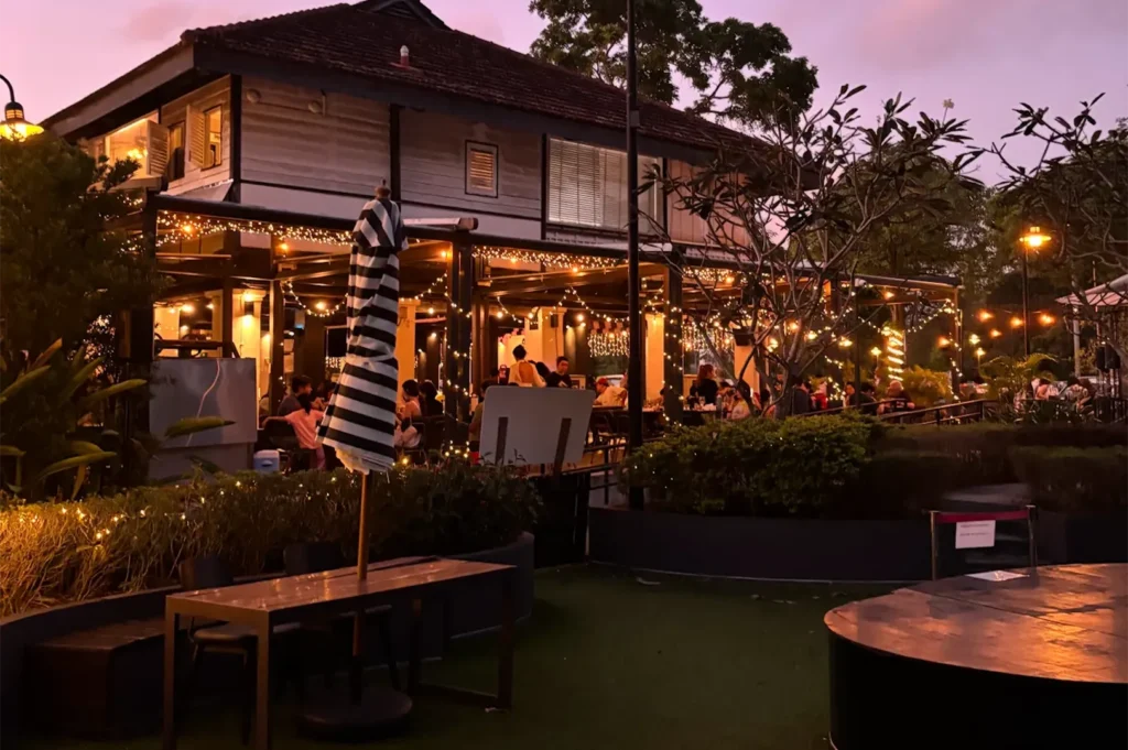 Scenic twilight view of an outdoor restaurant dining terrace crowded with patrons, lit by warm string lights under a beautiful purple evening sky.
