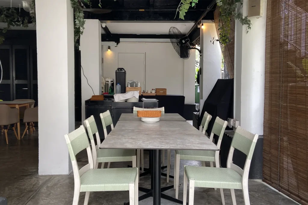 An empty long gray dining table arranged with light green woven chairs under a covered outdoor patio area featuring industrial fans and hanging green ivy.