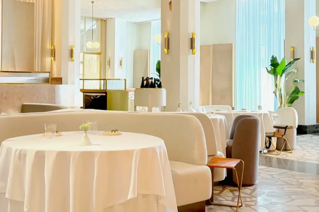 Bright and airy luxury restaurant interior with plush white booth seating, round table with white tablecloth, and tall indoor plants.