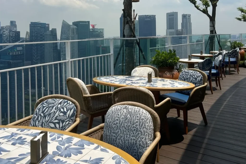 Outdoor rooftop restaurant terrace with patterned dining tables, woven chairs, and high-rise city skyscrapers in the background.