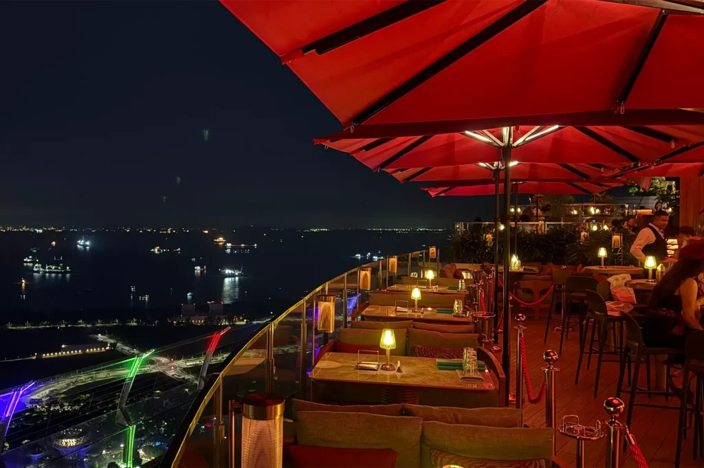 Nighttime rooftop lounge dining area under large red umbrellas with ambient table lamps looking out over the illuminated harbor.