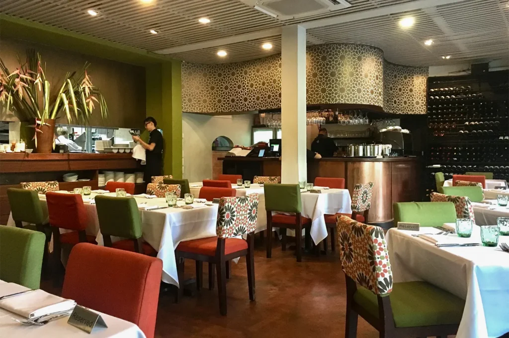 Warm and colorful restaurant interior with geometric mosaic wall, green and red upholstered dining chairs, and a wine display bar.