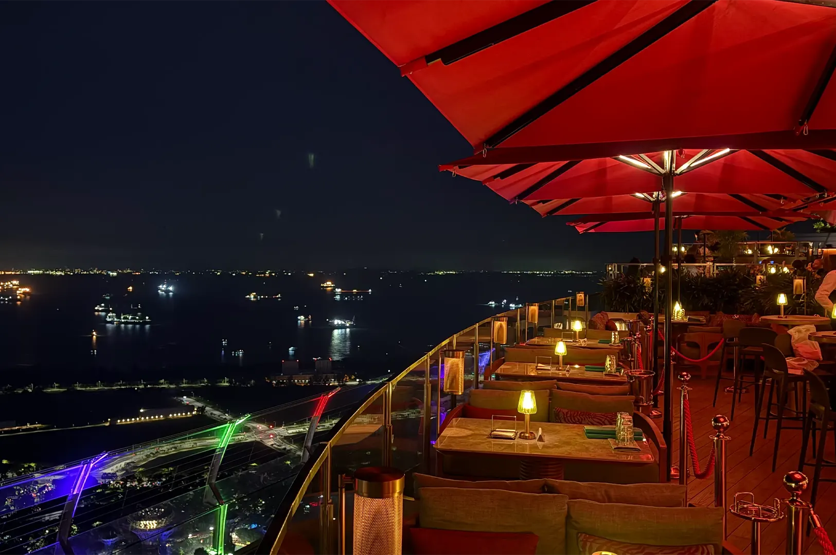 Outdoor rooftop dining terrace at CÉ LA VI Singapore overlooking the ocean and city lights at night under large red umbrellas.