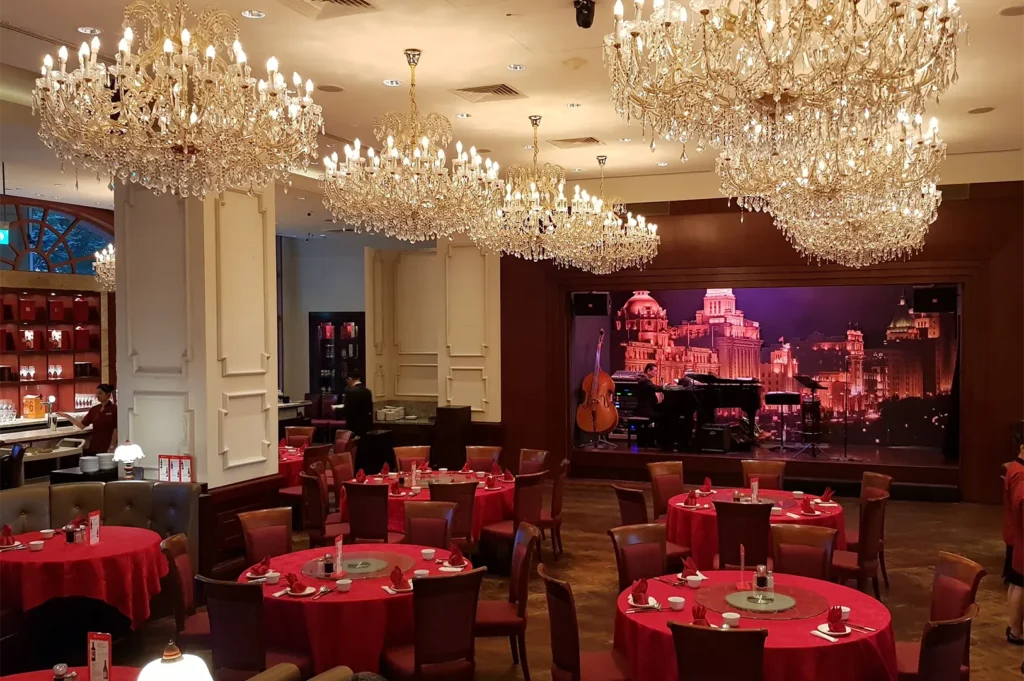 Luxurious dining hall at Grand Shanghai Restaurant featuring ornate crystal chandeliers, round tables with red tablecloths, and a live performance stage.