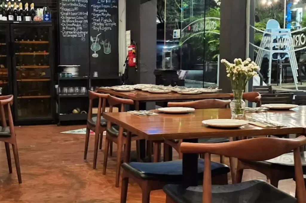 Cozy indoor dining area at Original Sin featuring wooden tables and a chalkboard menu displaying wine, beer, and mocktail selections.