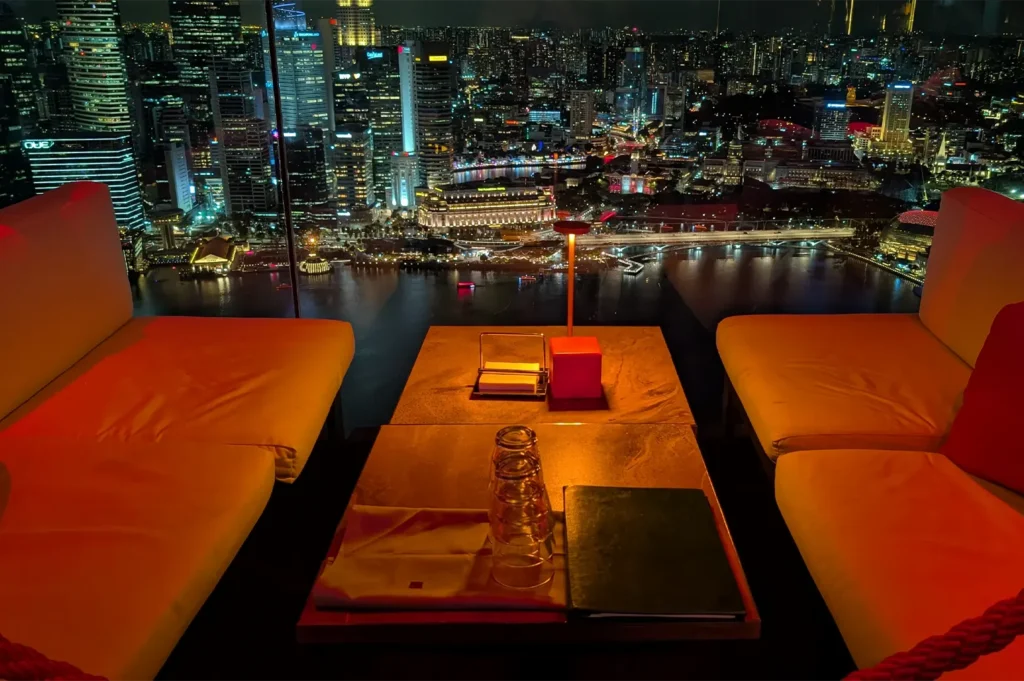Cozy rooftop sofa seating with warm ambient light overlooking the illuminated Singapore skyline and Marina Bay at night.