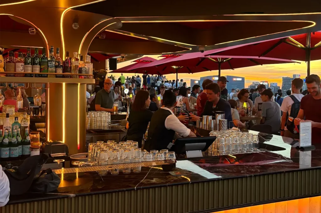 Crowded outdoor rooftop sky bar counter at CÉ LA VI with bartenders preparing drinks during sunset.