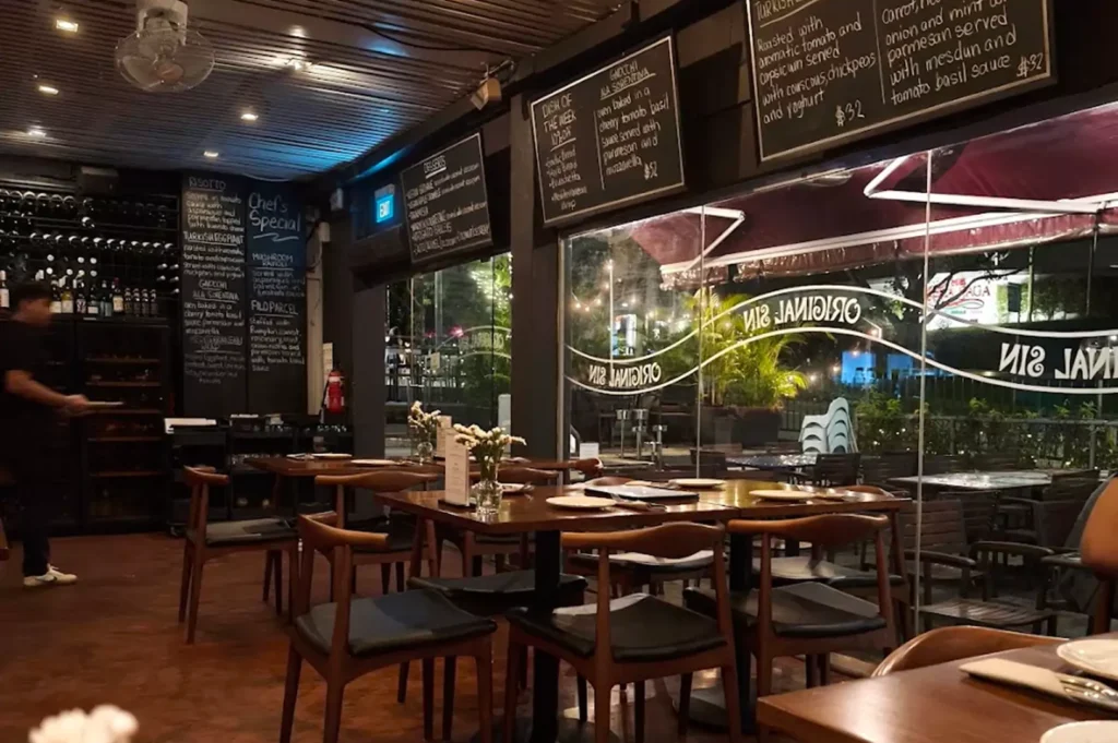 Spacious indoor dining room of Original Sin restaurant with wooden furniture, wine storage, and chalkboard listing Chef's Specials.