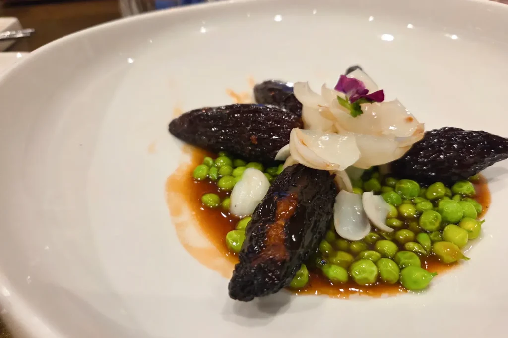 Gourmet braised sea cucumber served over a bed of green peas with delicate lily bulb garnish in a white bowl.