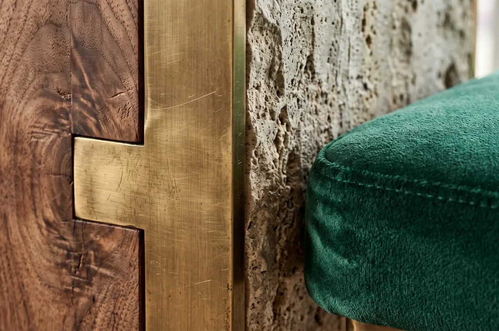 Macro detail shot showcasing premium interior design materials including dark walnut wood with a custom brass inlay joint, textured natural stone, and rich emerald green velvet upholstery.