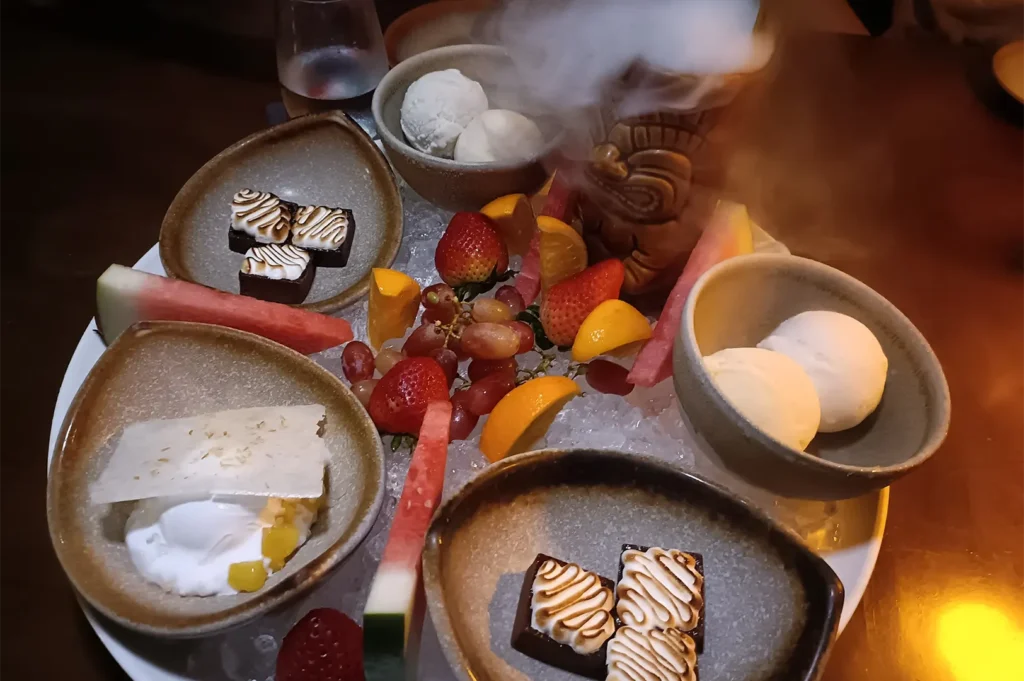 Gourmet dessert platter featuring fresh tropical fruits, toasted marshmallow bites, and scoops of ice cream with dry ice smoke effect.