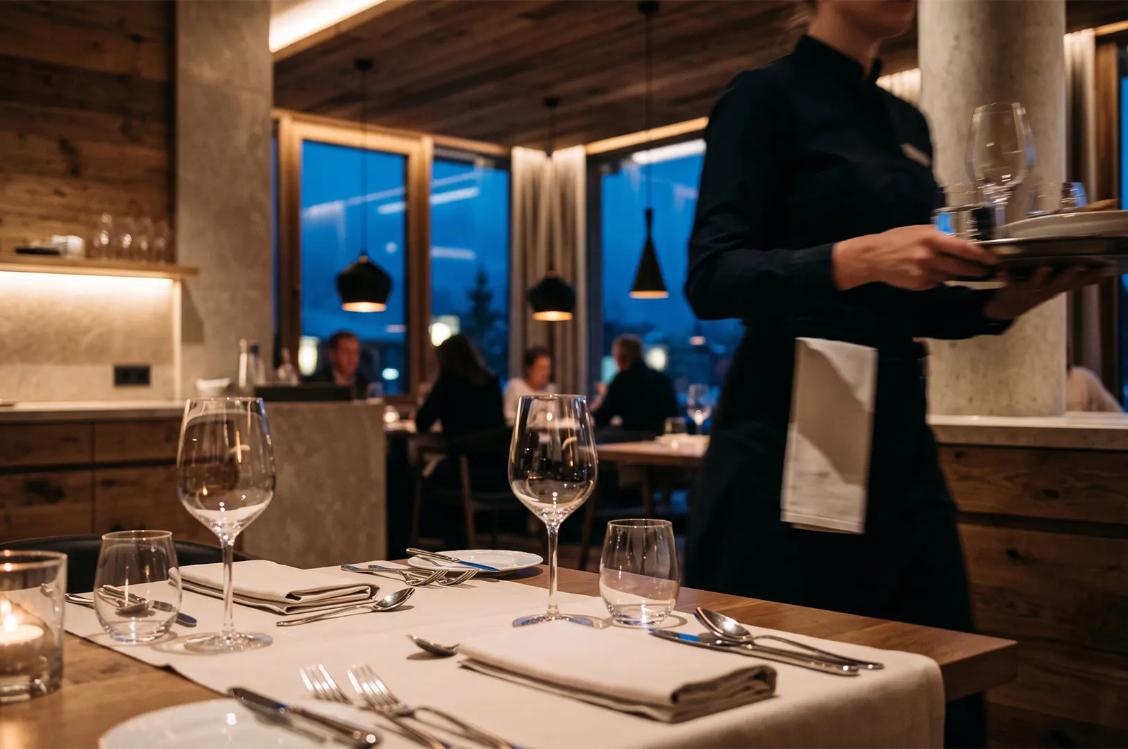Elegant fine dining table setting with wine glasses and cutlery, featuring a waitress carrying a tray in a dimly lit, atmospheric restaurant interior.