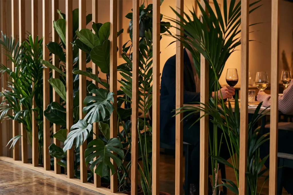 Guests enjoying wine behind a wooden slatted divider surrounded by green monstera and palm plants in a modern restaurant interior.