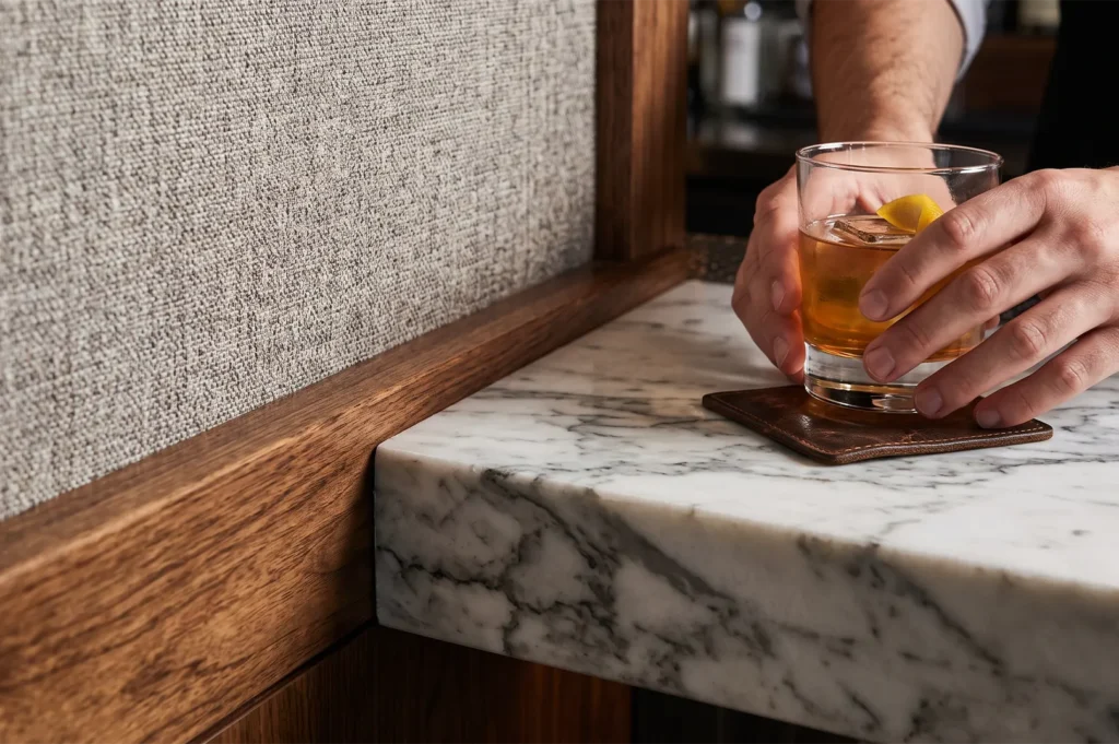 Bartender placing a craft whiskey cocktail with a lemon peel garnish on a leather coaster atop a white marble bar counter.